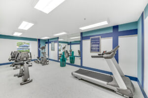 Interior Fitness Center, padded floor, treadmill, elliptical machine , stationary bikes, mirrored wall, basket with yoga mats, blue, cyan and white walls.