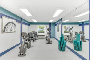 Interior Fitness Center, padded floor, treadmill, elliptical machine , stationary bikes, mirrored wall, basket with yoga mats, blue, cyan and white walls, inspirational quote on wall.