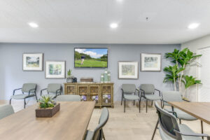 Interior Community Room, Square tables, wood floor, tv mounted on wall, light blue walls, landscape paintings on walls, succulents on tables.