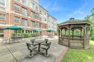 Exterior Vistas at Lake Largo Apartments, patio/ gazebo area, bistro tables with umbrellas, red brick and beige building with white trim in background, natural wood gazebo, photo taken on a sunny day with clouds.