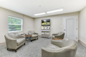 Interior community room, lounge chairs, beige walls, neutral toned carpeting, end tables, tv mounted to wall.