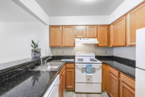 Interior unit kitchen, stainless steel sink, white appliances, black stone countertop, light brown wood cabinets, wood floors.