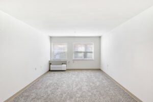 Interior Living Room, neutral toned carpeting, white walls, 2 windows electrical sockets, ac unit in corner of the room.