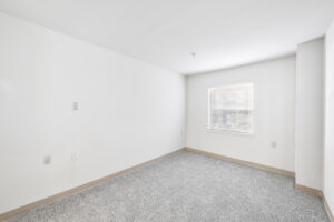 Interior Unit Bedroom, neutral toned carpeting, white walls, electrical sockets, window.
